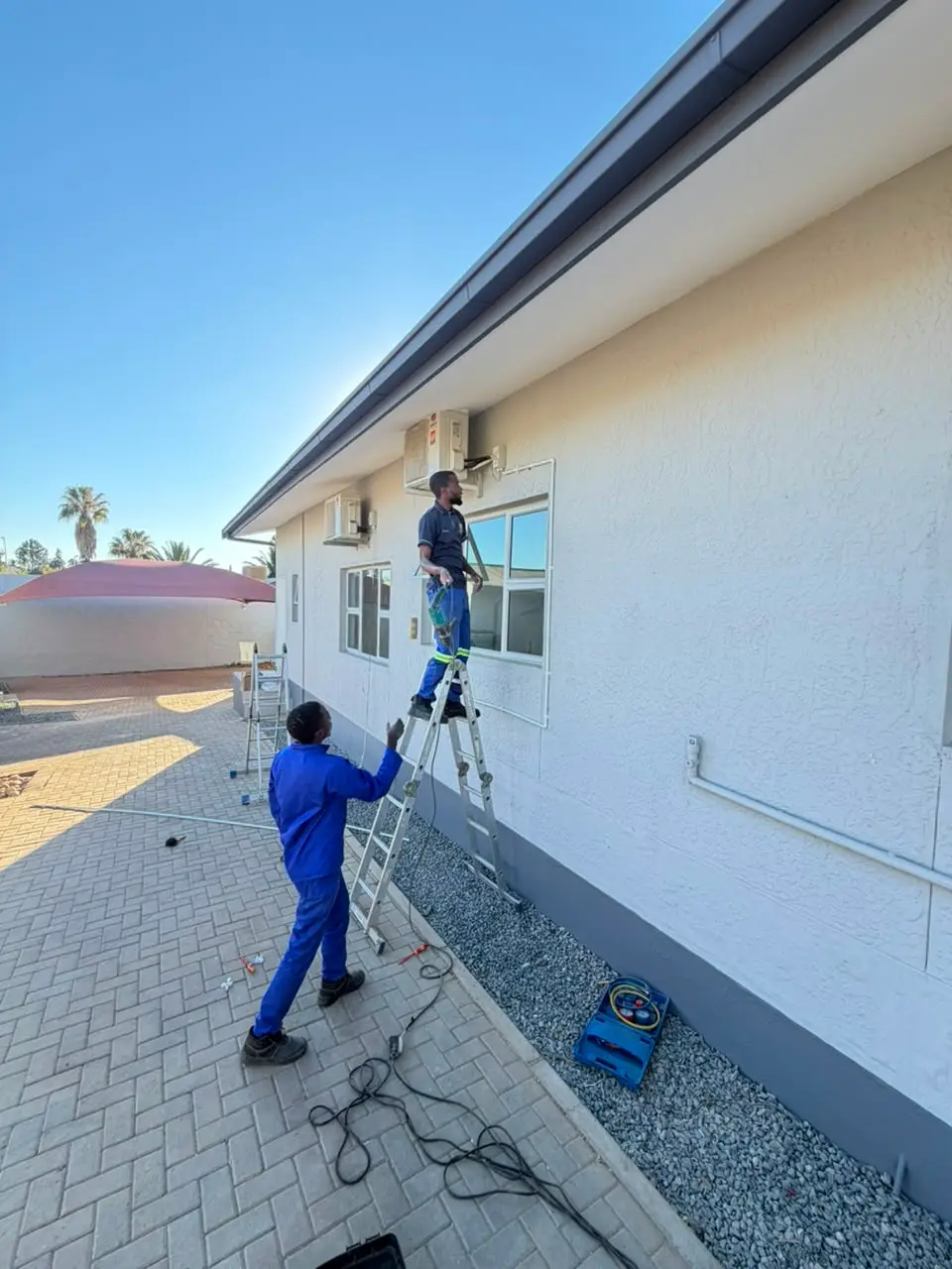 Two technicians installing split air conditioning units on a building wall
